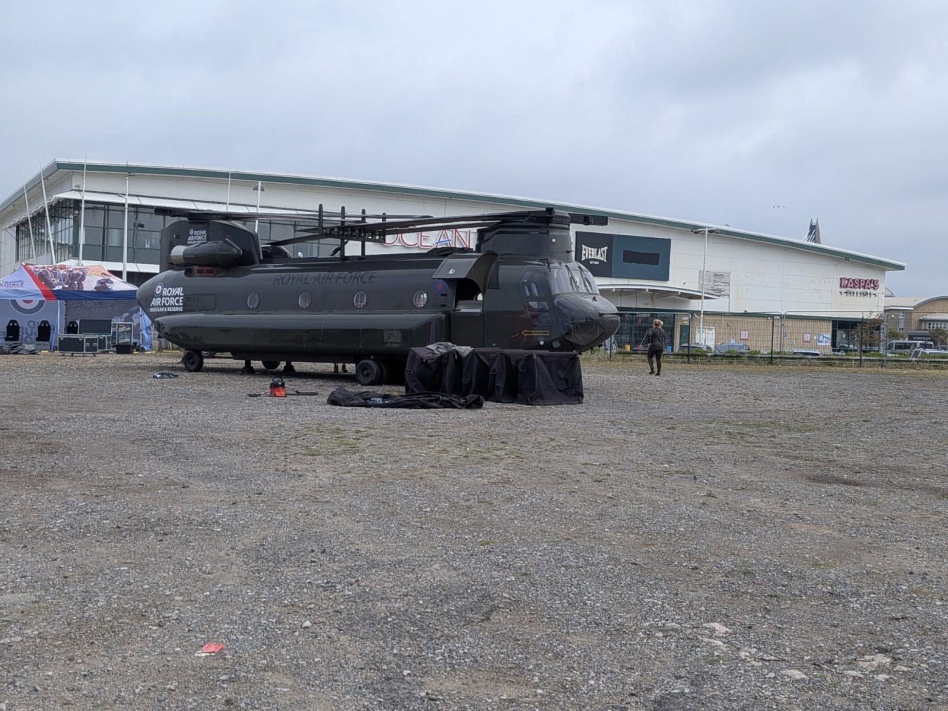 Military helicopter on a tarmac outside a building with banners and tents nearby.