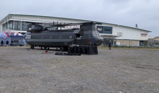 Military helicopter on a tarmac outside a building with banners and tents nearby.