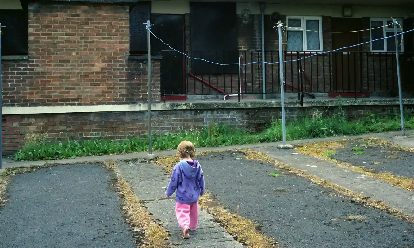 Child wearing purple jacket walks away from camera along a cracked, leaf-strewn pavement beside a brick building with a railing and clotheslines.