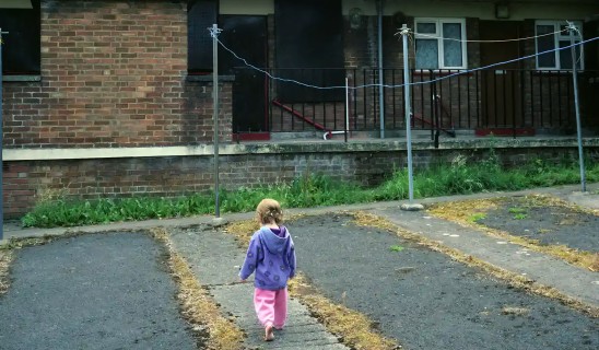 Child wearing purple jacket walks away from camera along a cracked, leaf-strewn pavement beside a brick building with a railing and clotheslines.