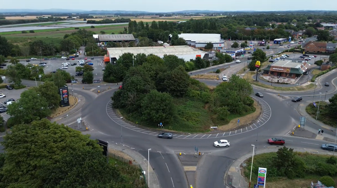 Aerial view of a roundabout with surrounding shops, cars, and green trees.