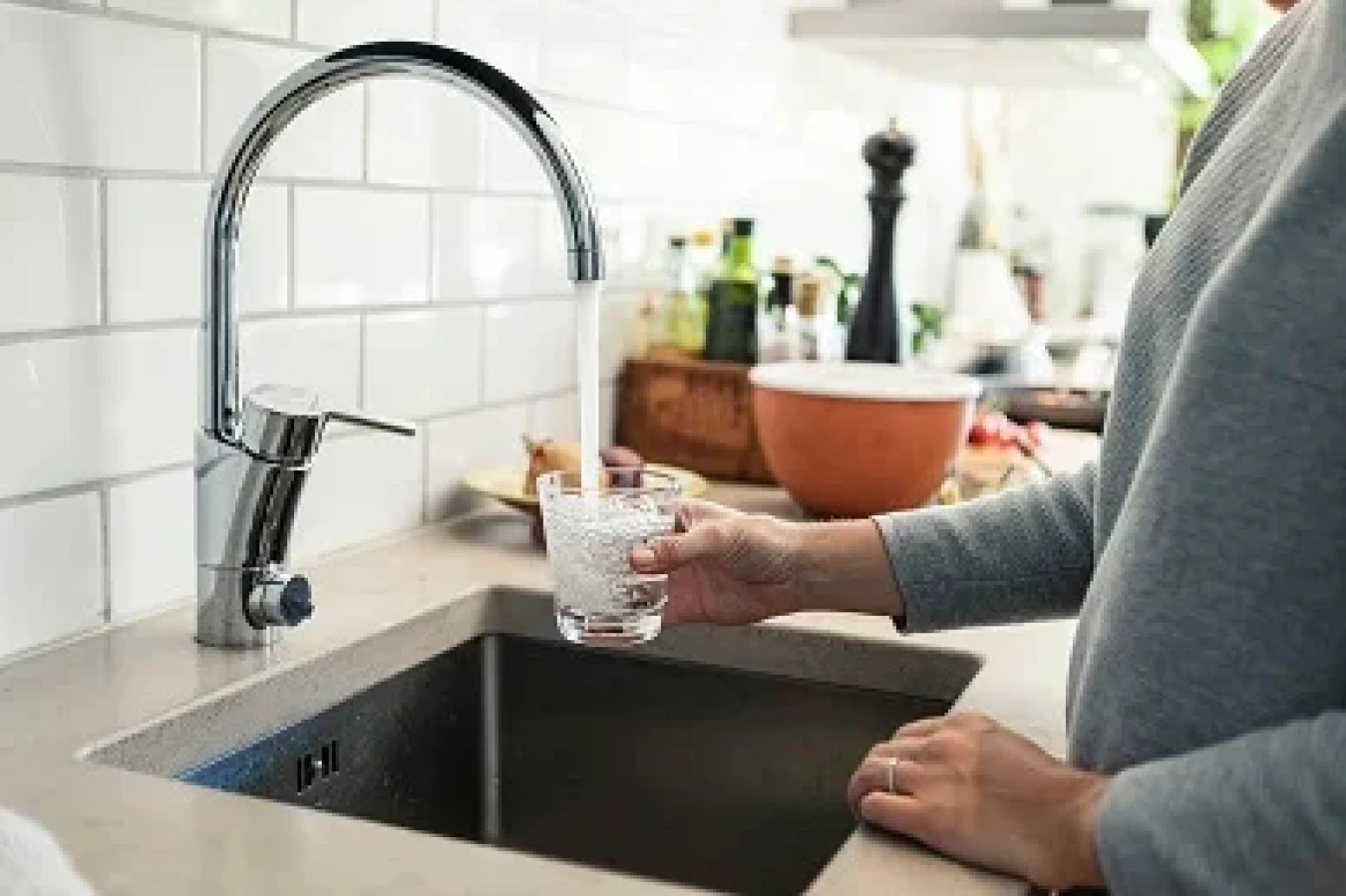 A person rinses a glass at a modern kitchen sink with chrome faucet and white subway tile backsplash.