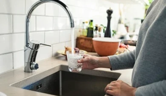 A person rinses a glass at a modern kitchen sink with chrome faucet and white subway tile backsplash.
