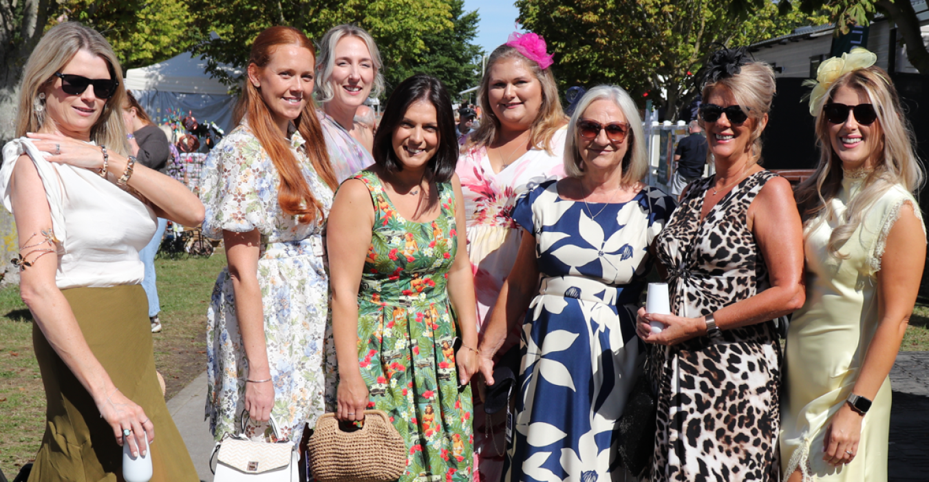 Group of women posing outdoors in summer dresses, smiling, with tents and trees in background.