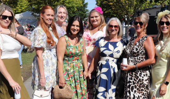 Group of women posing outdoors in summer dresses, smiling, with tents and trees in background.