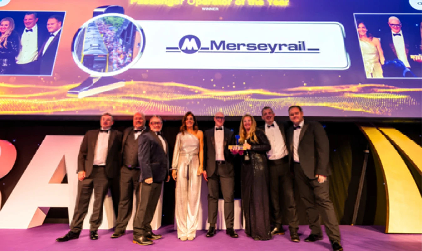 Group of formally dressed people posing on stage at an event with a large screen and signage in the background