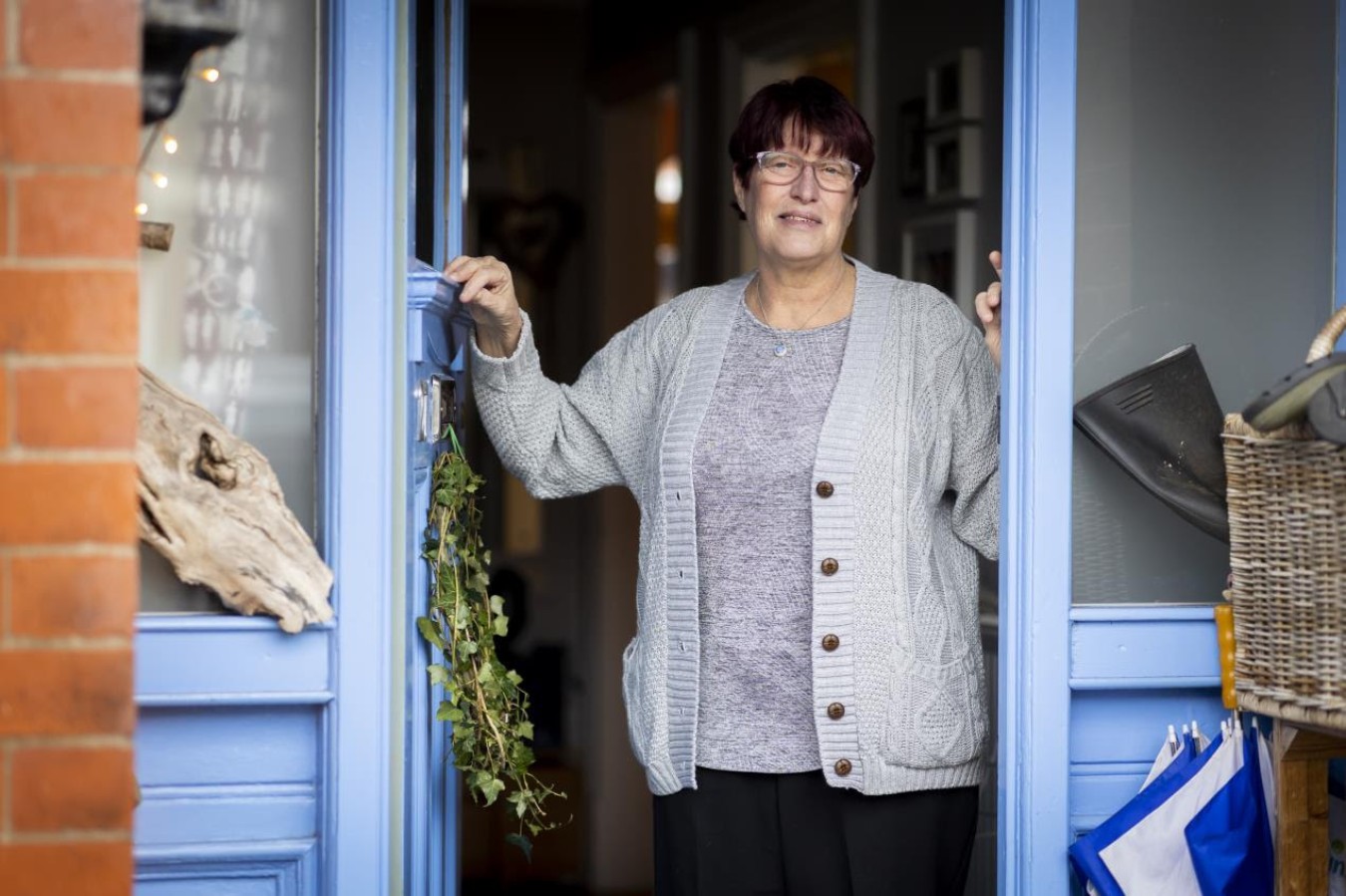 woman standing in doorway of blue-painted shop, smiling, wearing glasses and a grey cardigan