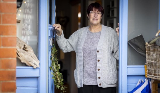 woman standing in doorway of blue-painted shop, smiling, wearing glasses and a grey cardigan