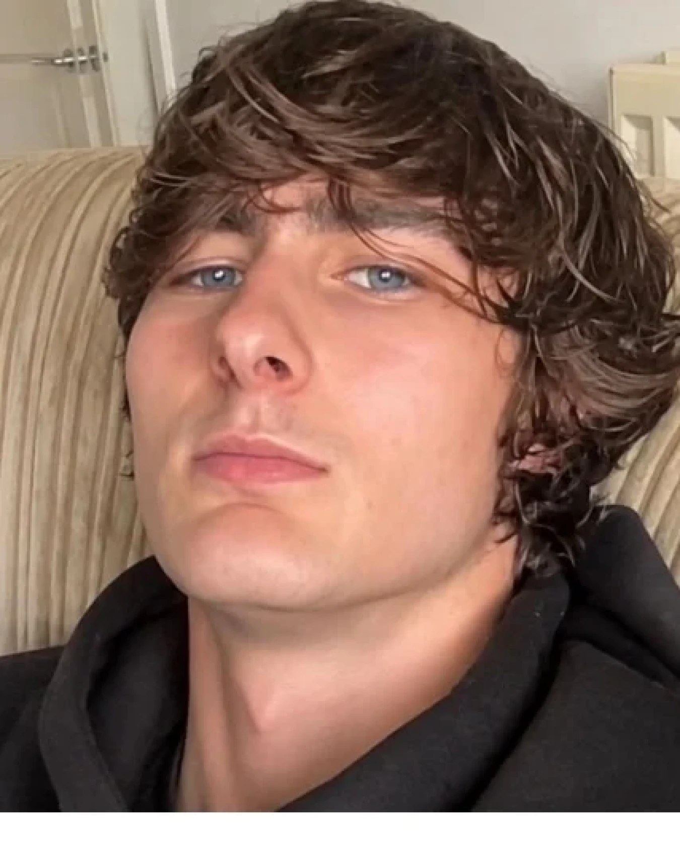 young man with wavy brown hair sitting on a sofa, wearing a dark hoodie