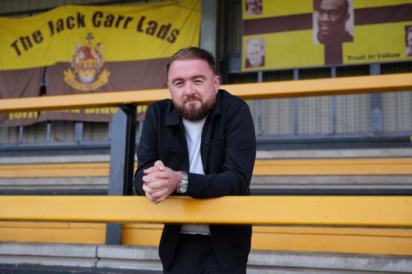 Man with beard leaning on yellow railing in stadium stands, banners in background reading The Jack Carr Lads.