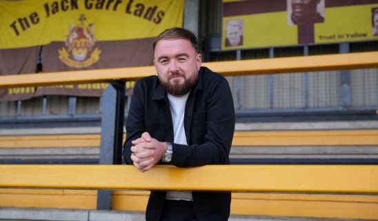Man with beard leaning on yellow railing in stadium stands, banners in background reading The Jack Carr Lads.
