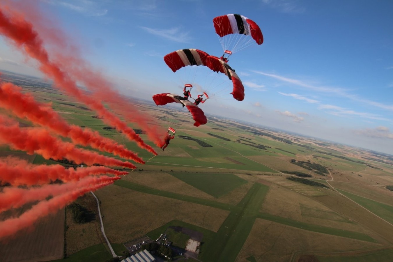 Skydivers deploy red-and-white parachutes above a patchwork countryside, leaving red smoke trails.