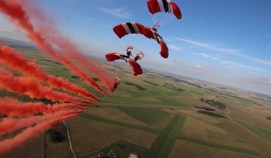 Skydivers deploy red-and-white parachutes above a patchwork countryside, leaving red smoke trails.