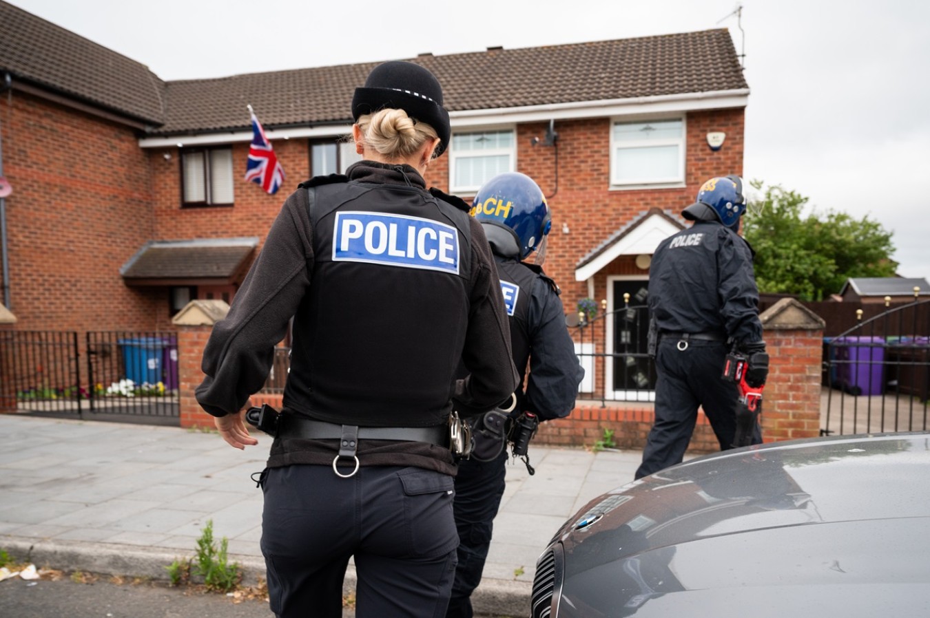 Police officers in protective gear on a residential street near a brick house with a Union Flag