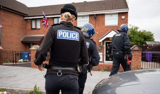 Police officers in protective gear on a residential street near a brick house with a Union Flag
