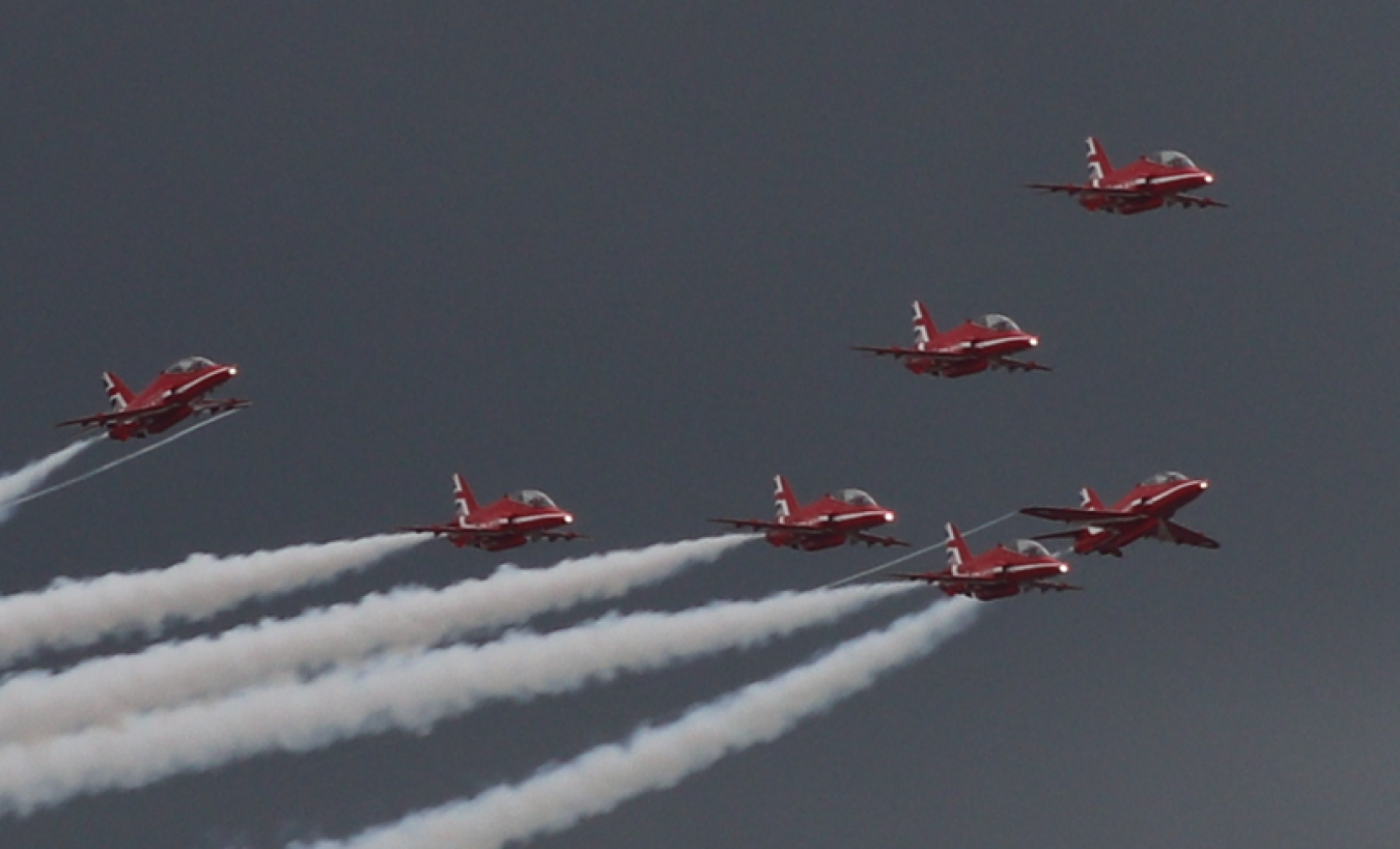 Six red aerobatic aircraft in formation flight against a dark sky with white smoke trails