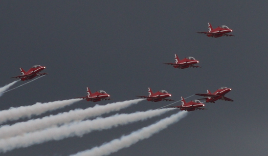 Six red aerobatic aircraft in formation flight against a dark sky with white smoke trails