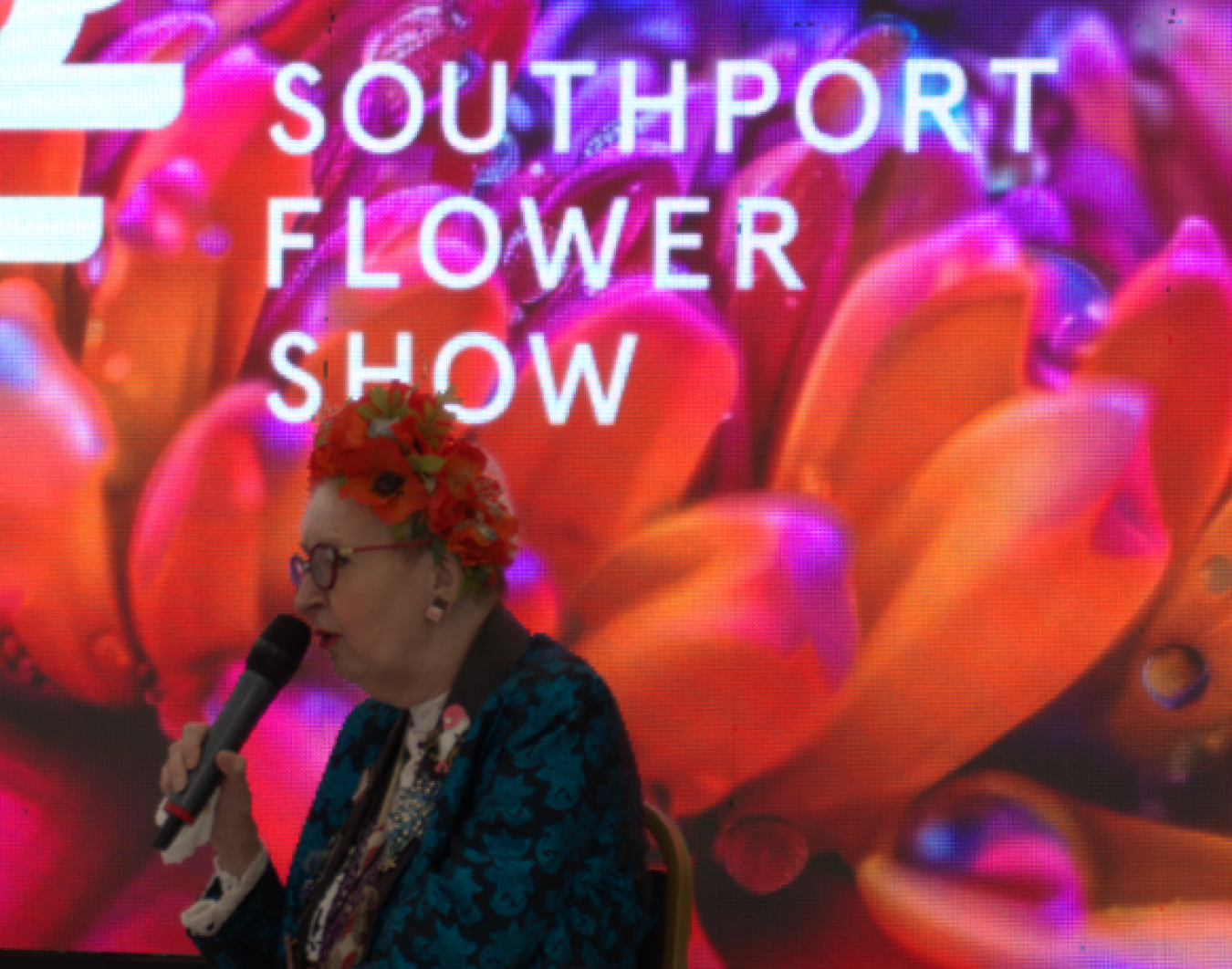 Elderly woman speaking into microphone at event with vibrant floral backdrop reading Southport Flower Show.