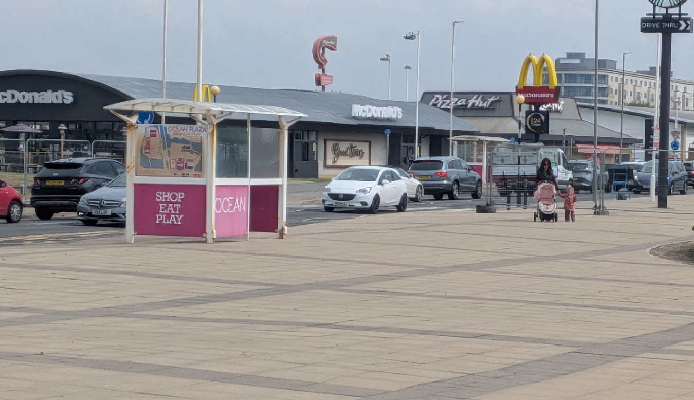 Mall frontage with McDonald's, Pizza Hut and Ocean Plaza shop units; a white car and other vehicles parked, with a small pink kiosk in the foreground.