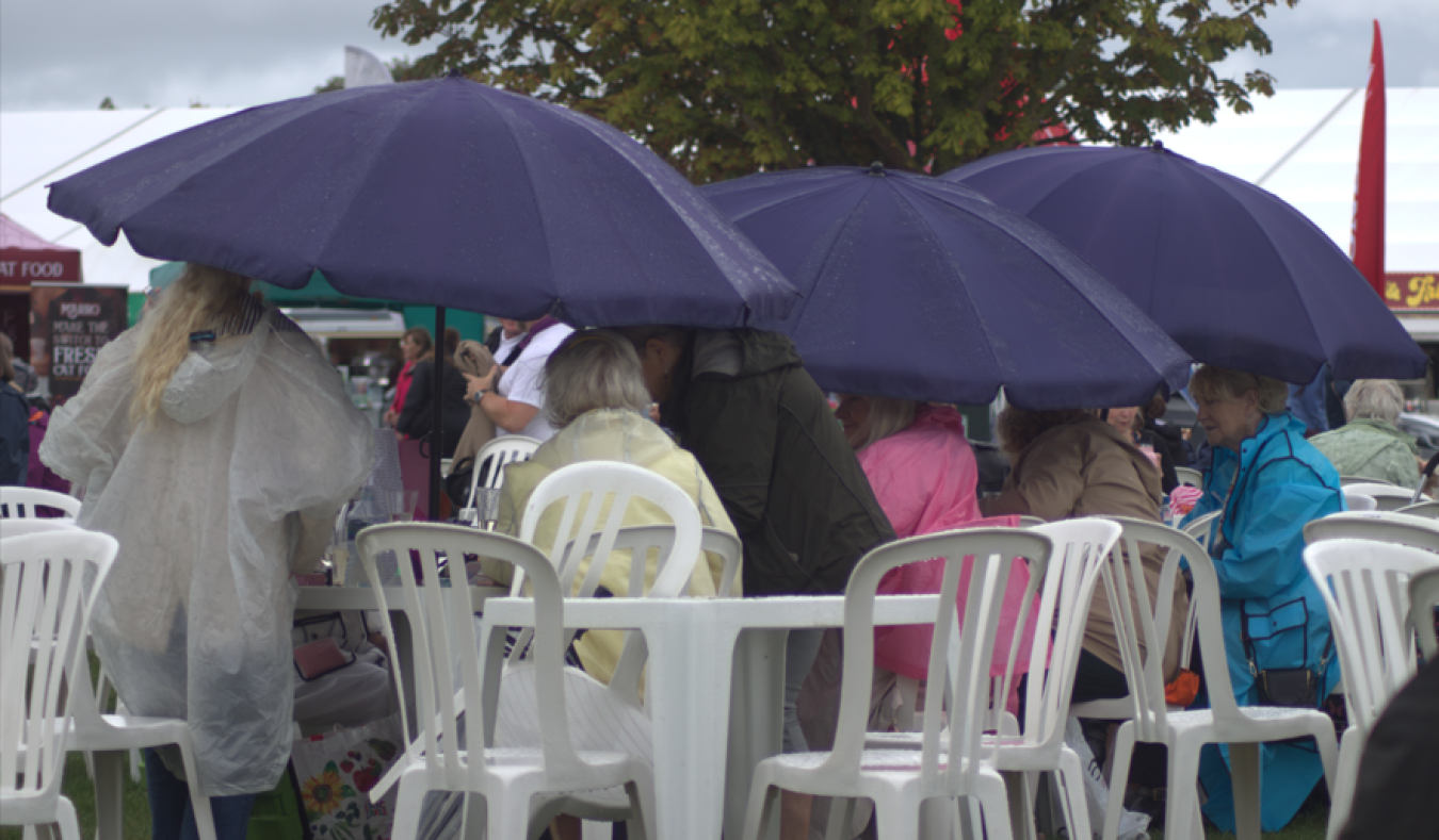 People seated under large blue umbrellas at an outdoor event with plastic chairs.