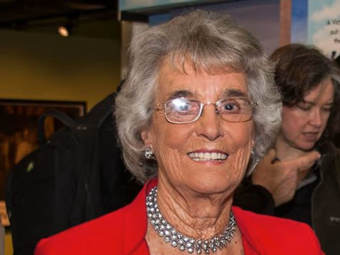 Smiling elderly woman with short grey hair and glasses wearing a red outfit and a beaded necklace.
