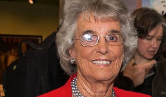 Smiling elderly woman with short grey hair and glasses wearing a red outfit and a beaded necklace.