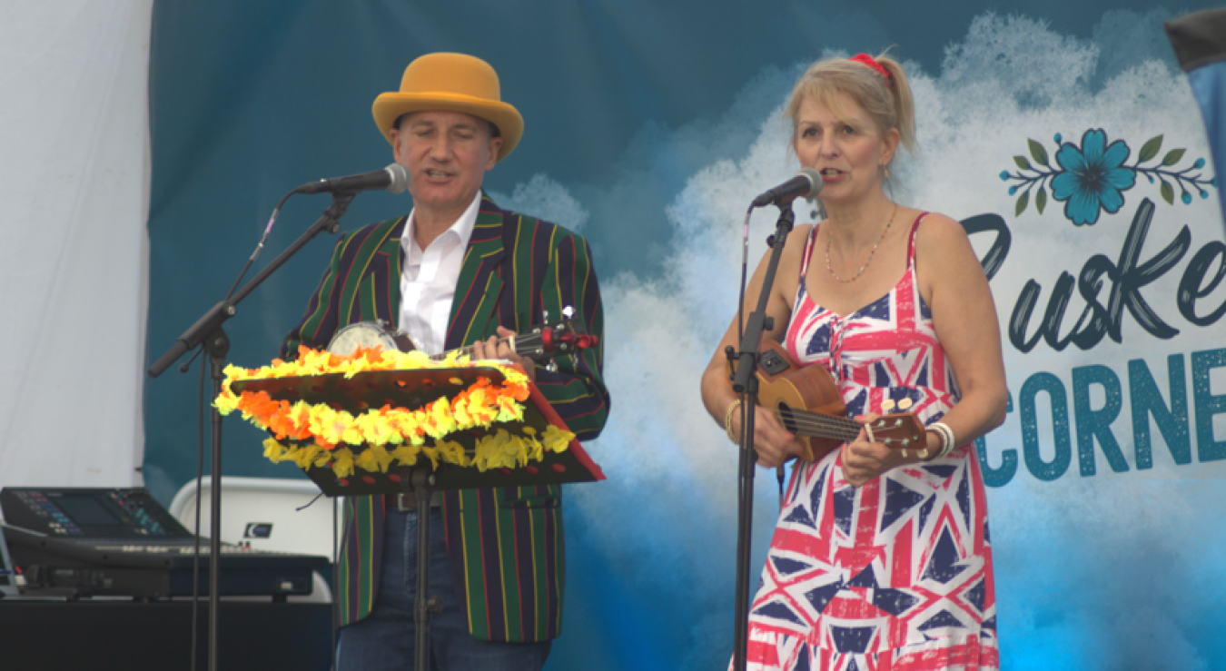 Two performers on stage, man in striped jacket and tan hat, woman in Union Jack dress with ukulele, microphones and flower decoration on lectern.