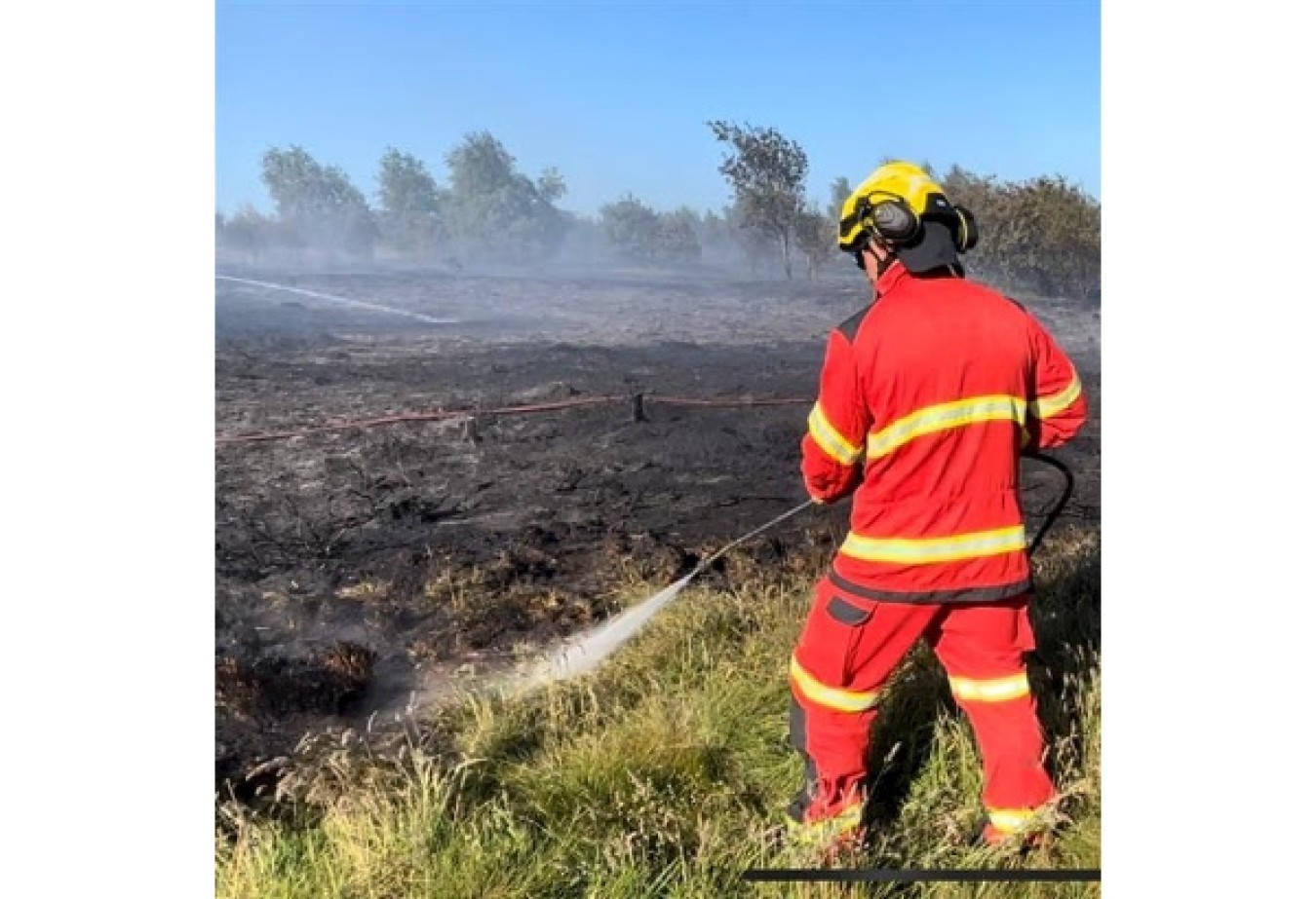 Firefighter in red protective gear hosing a grassland fire on a sunny day