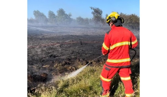 Firefighter in red protective gear hosing a grassland fire on a sunny day