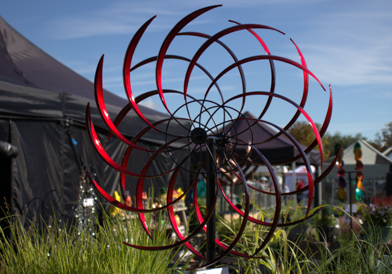 Red spiral metal sculpture in garden near tents at outdoor fair.