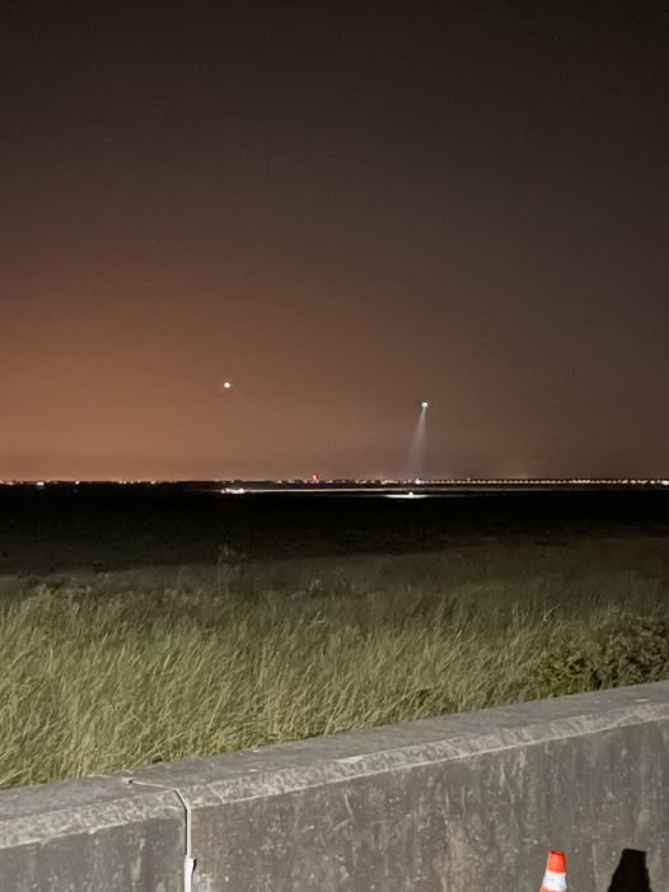 Nighttime landscape with distant lights and a spotlight piercing the sky, low grassy foreground and a concrete barrier in the front right