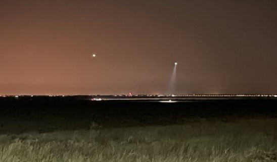 Nighttime landscape with distant lights and a spotlight piercing the sky, low grassy foreground and a concrete barrier in the front right