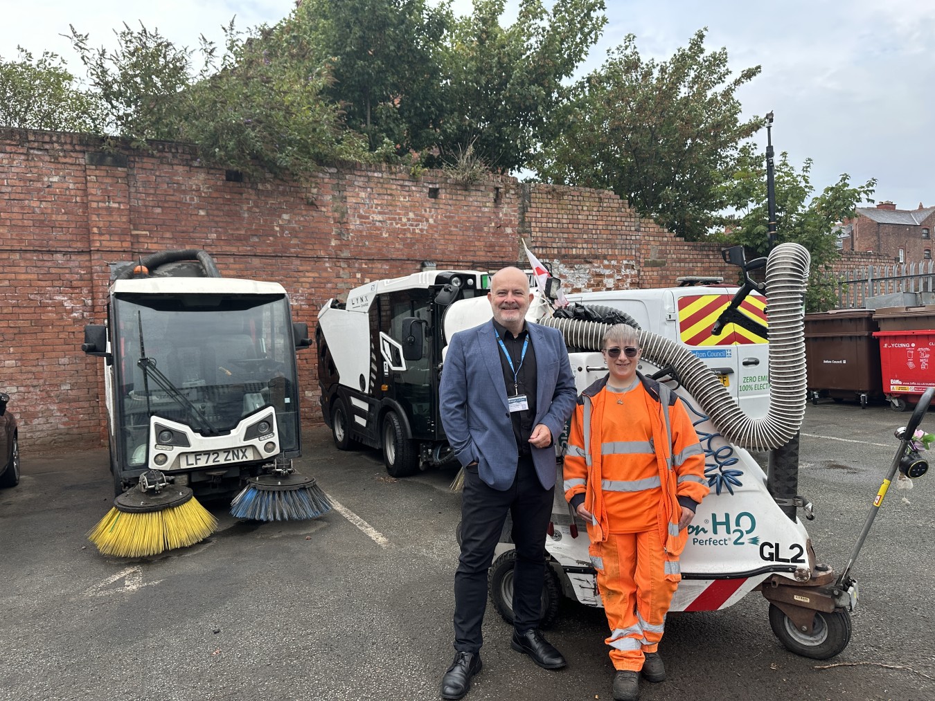 Two people stand in a cluttered lot with street cleaning equipment and a red brick wall behind.