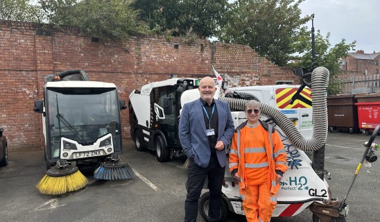 Two people stand in a cluttered lot with street cleaning equipment and a red brick wall behind.