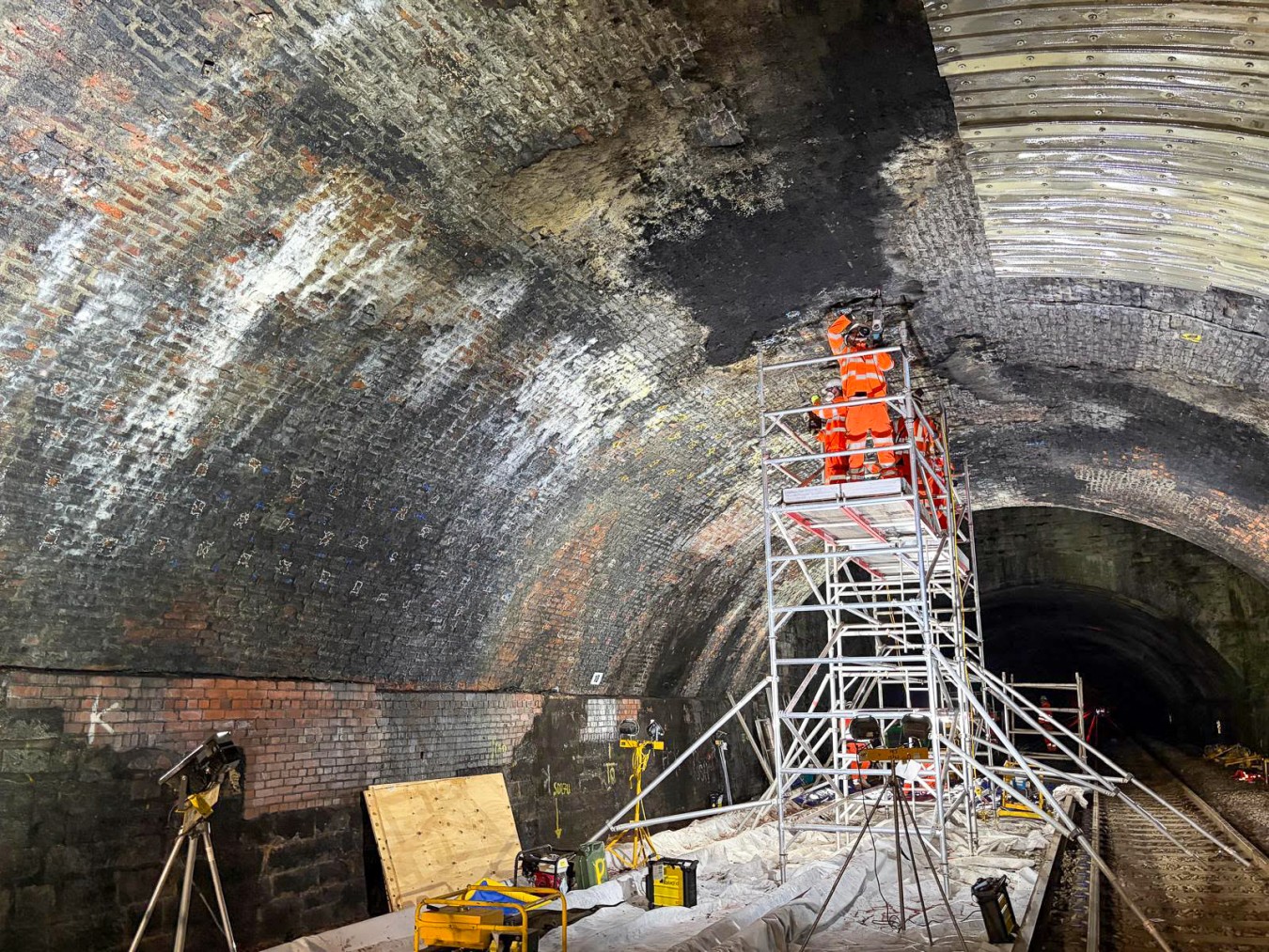 Workers in bright orange high-visibility clothing on scaffolding inside a curved brick tunnel.