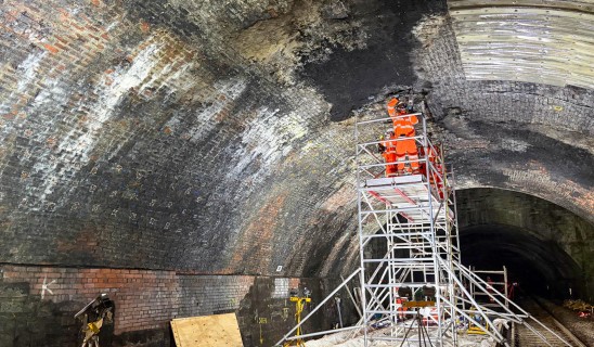 Workers in bright orange high-visibility clothing on scaffolding inside a curved brick tunnel.
