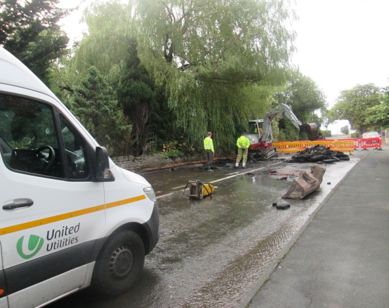 Road construction site with excavation machinery and barriers, temporary water on road, white van in foreground