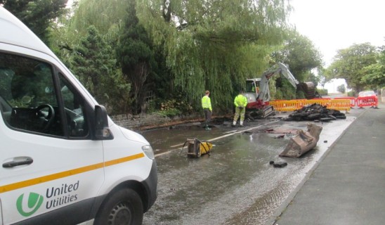 Road construction site with excavation machinery and barriers, temporary water on road, white van in foreground