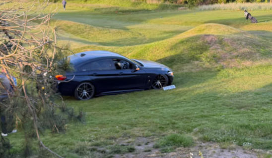 Sports car off the course on a grassy bank adjacent to undulating putting greens.