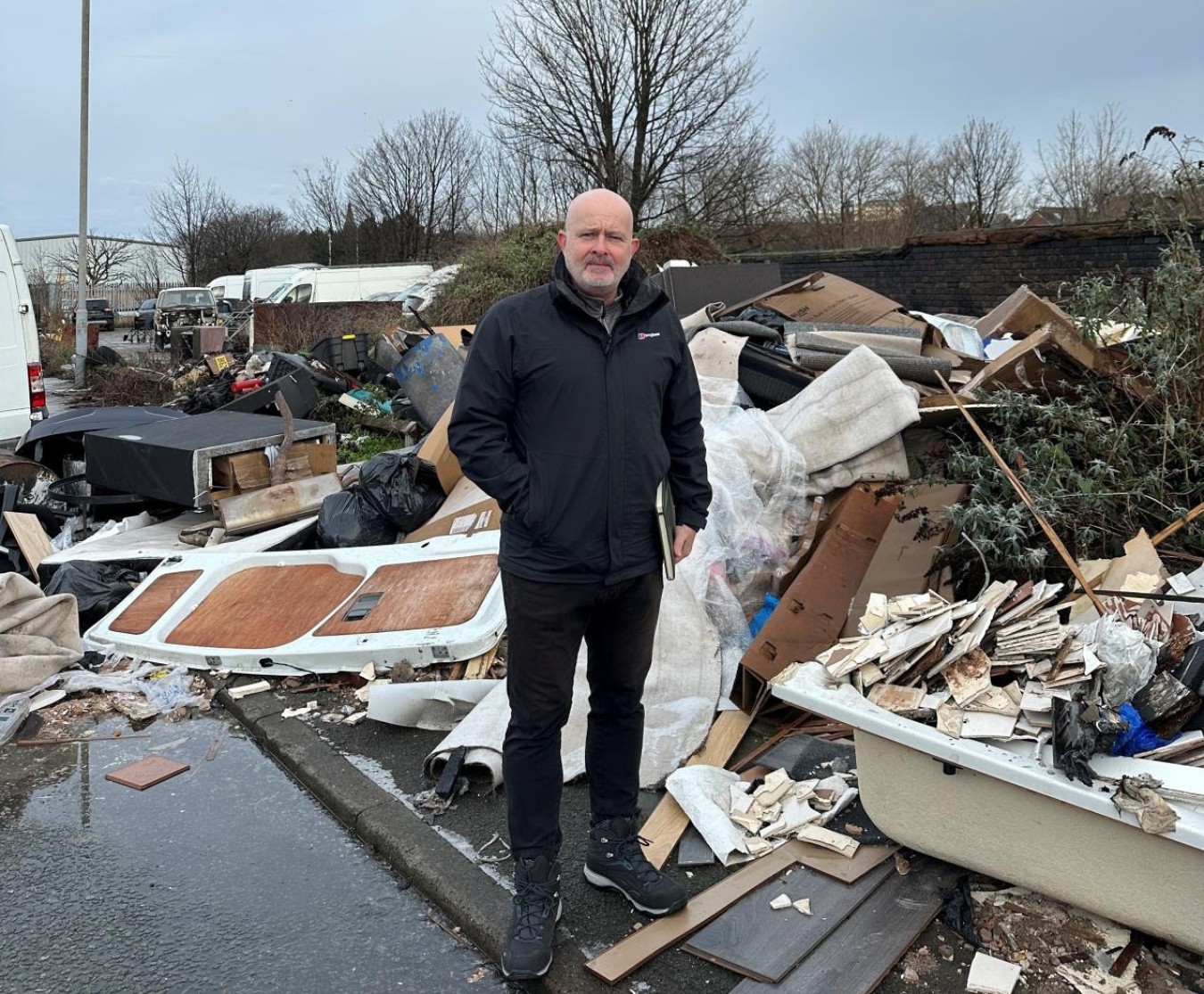 Bald man in black jacket stands among a heap of discarded debris and rubbish by a street.