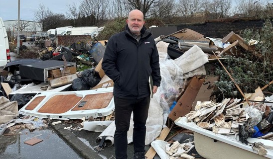Bald man in black jacket stands among a heap of discarded debris and rubbish by a street.
