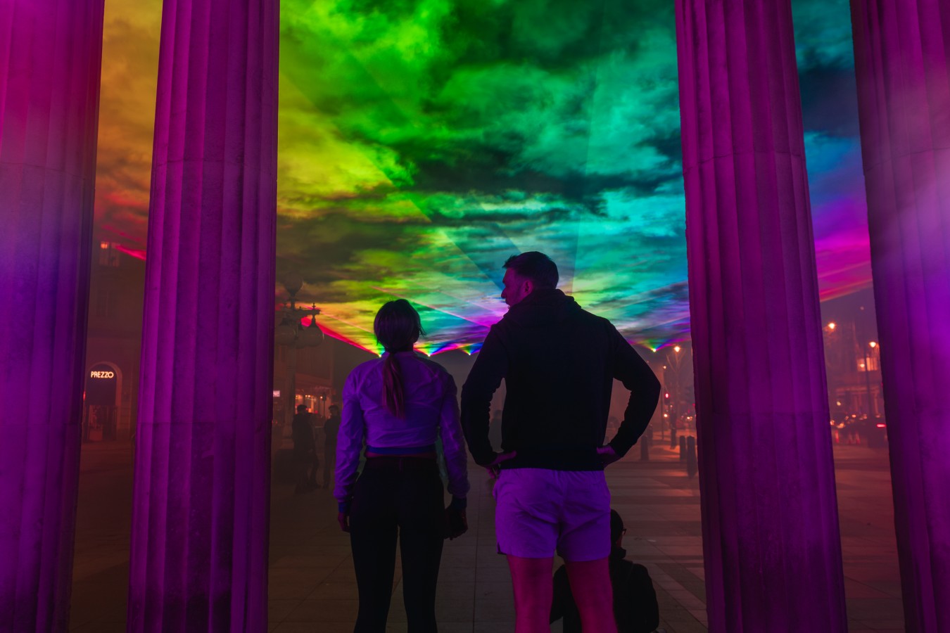 People stand among tall columns immersed in vibrant rainbow light projections.