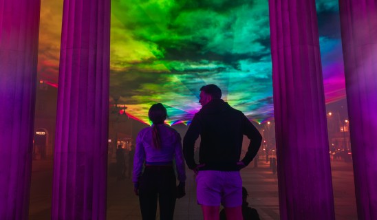 People stand among tall columns immersed in vibrant rainbow light projections.