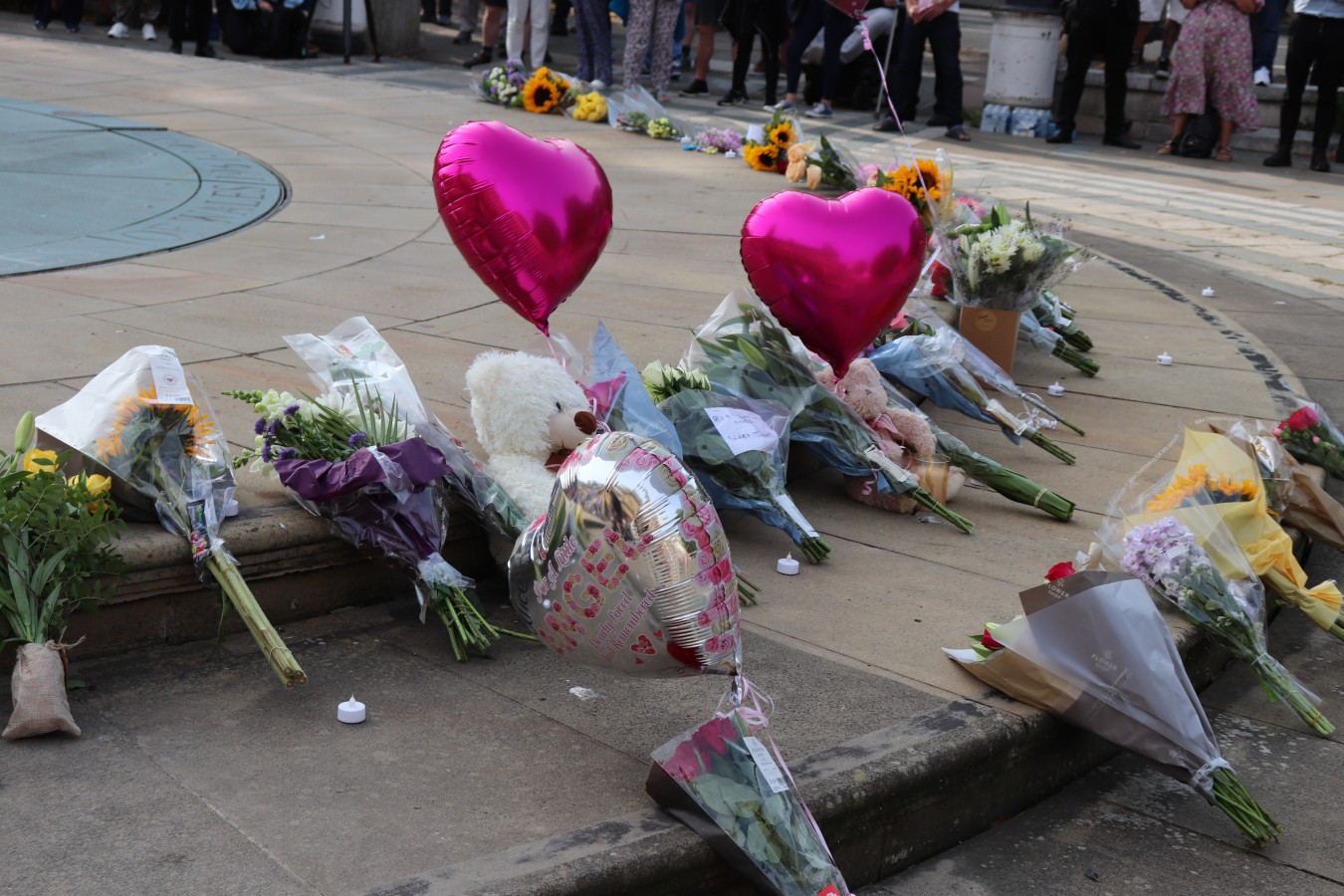 Memorial flowers, bouquets and balloons laid along a city pavement with a teddy bear and heart-shaped balloons
