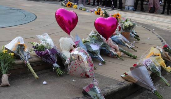 Memorial flowers, bouquets and balloons laid along a city pavement with a teddy bear and heart-shaped balloons