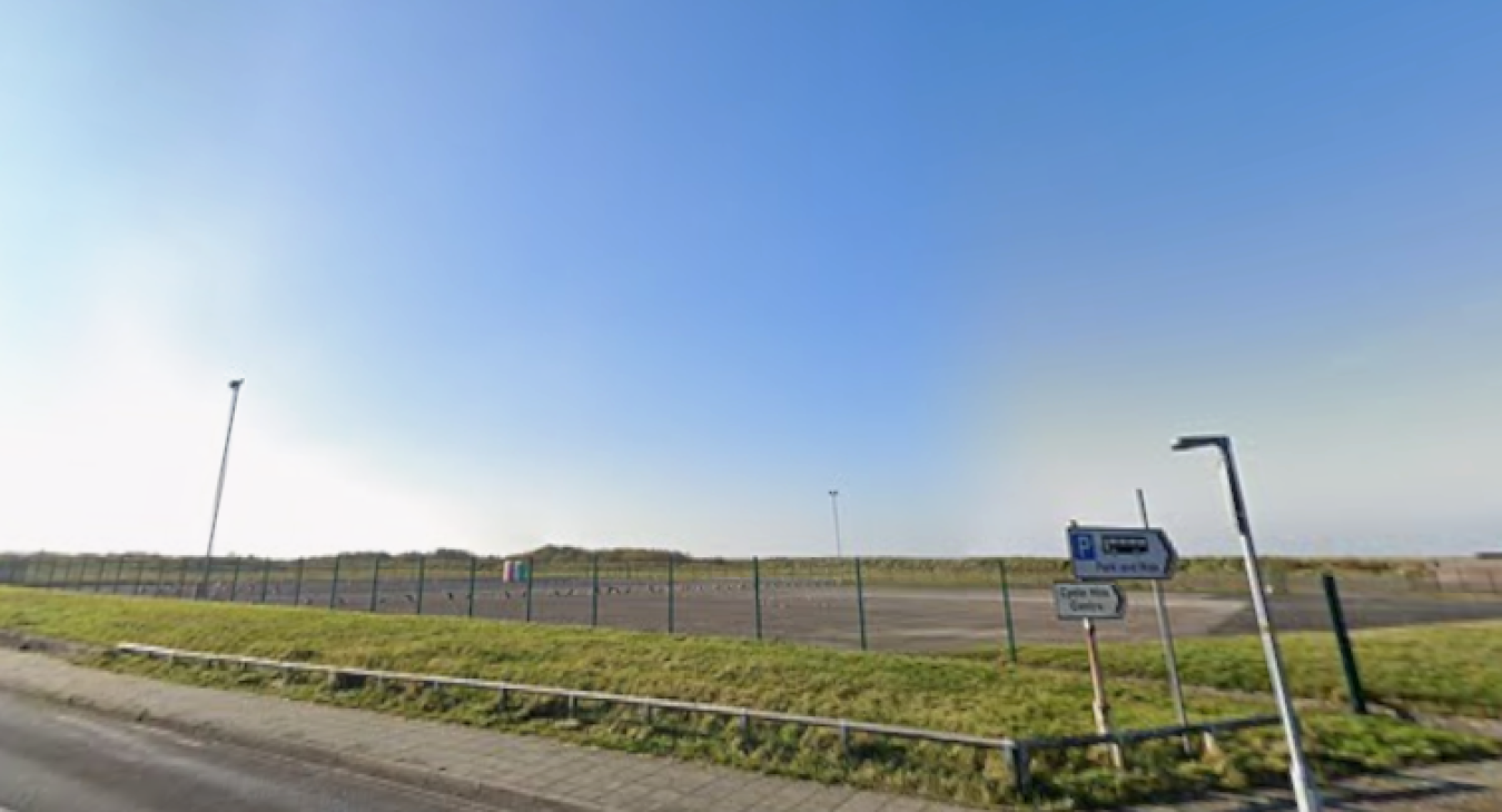 Open roadside area with fenced field, blue sky and a few street signs near a road.