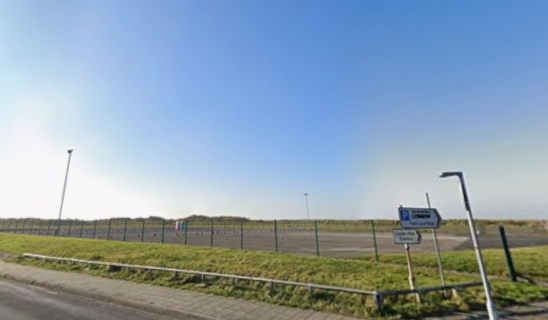Open roadside area with fenced field, blue sky and a few street signs near a road.