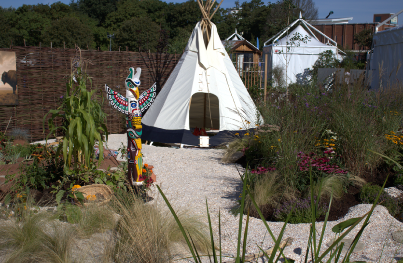A white tipi set in a garden with ornamental plants and a totem pole sculpture in the foreground