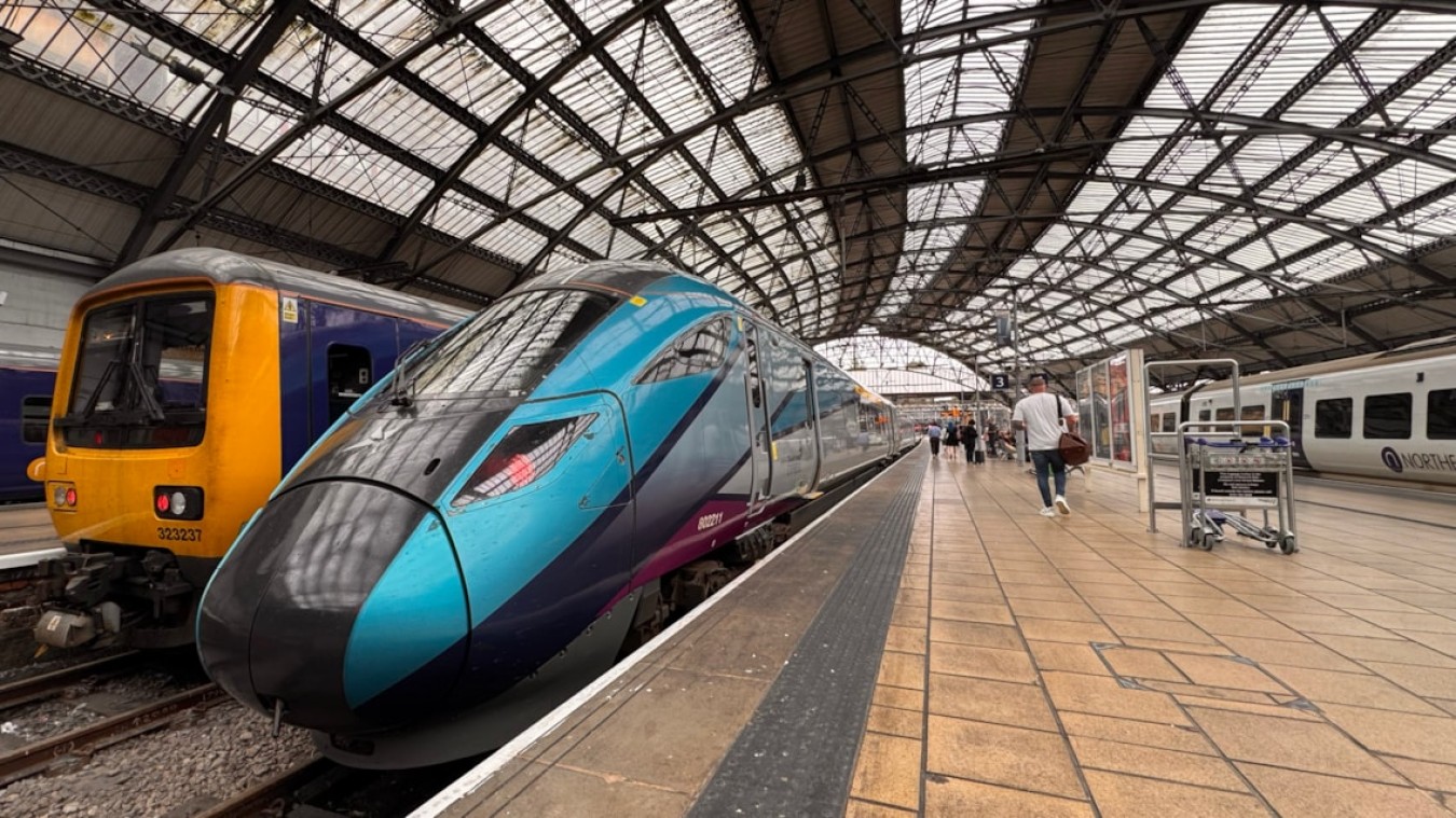 Modern blue and purple high-speed train at a station platform, with a yellow diesel train on adjacent track under a large arched glass roof.