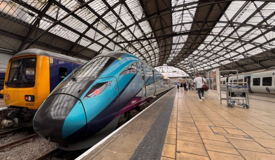 Modern blue and purple high-speed train at a station platform, with a yellow diesel train on adjacent track under a large arched glass roof.